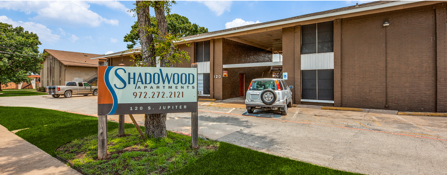 A driveway leads into Shadowood Apartments, with signage in view and the brick exterior building in the back.