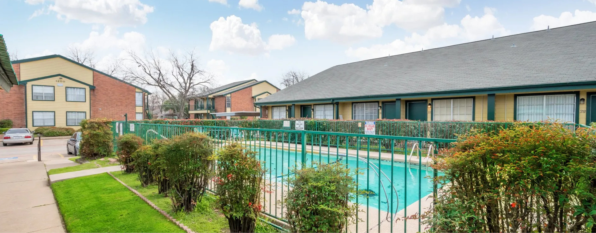 Brick and wood-paneled multifamily apartment, featuring a fenced outdoor pool and landscaped garden area.