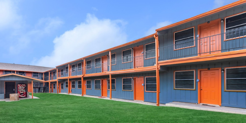 Vibrant two-story multifamily housing with orange doors and trim, gray siding, and a communal lawn area under a partly cloudy sky.