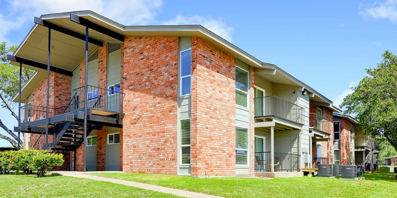 Brick multifamily apartment complex with external metal staircases, upper-level balconies, and well-maintained lawns under a clear blue sky.