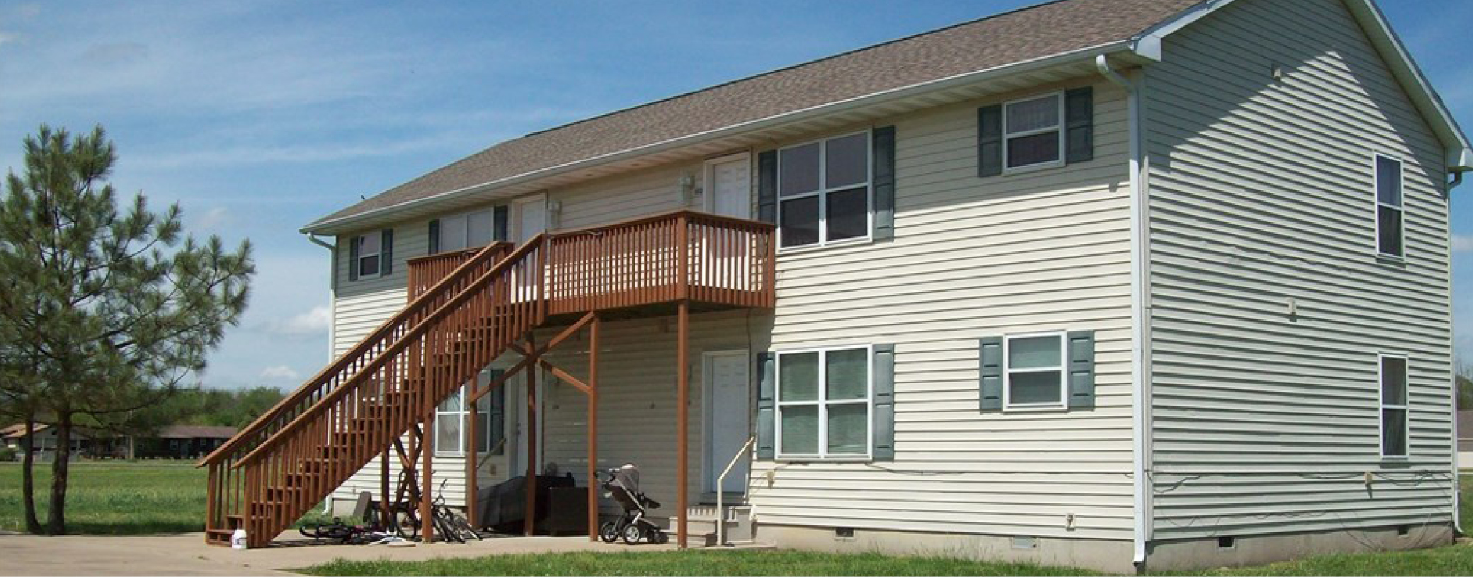 Two-story multifamily building with beige siding and wooden staircases leading to upper-level units, set against a backdrop of clear skies and greenery.
