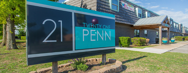 Sign for 'TWENTY ONE PENN' in front of the apartment complex with modern design elements, landscaped beds, and a clear blue sky.
