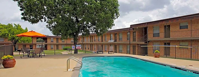 Outdoor swimming pool area with patio furniture and an orange umbrella, surrounded by a two-story brick apartment building, under a cloudy sky.