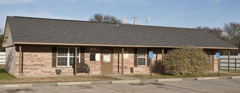 A single-story brick building with numbered doors, accessible parking signs, and a wooden bench, under a clear sky.