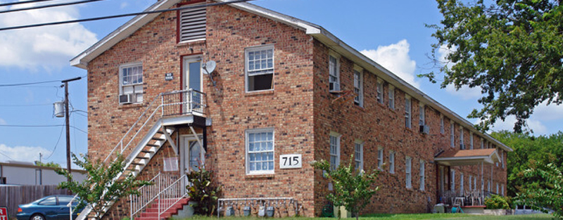 A two-story red brick apartment building with exterior stairs, number 715 displayed, and a blue car parked on the side, under a partly cloudy sky.