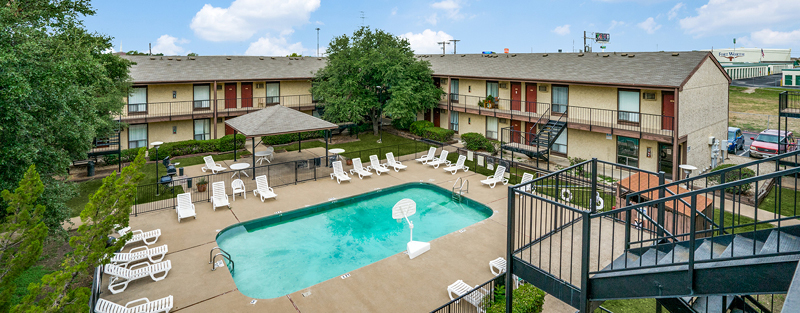 Outdoor pool with chairs and a shaded area, encircled by a two-story apartment complex, on a day with a cloudy sky.