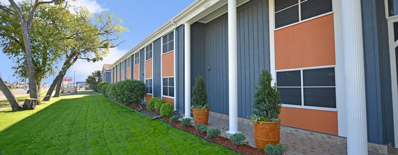 Long two-story apartment building with orange and blue facade, white columns, and landscaped lawn with potted plants.
