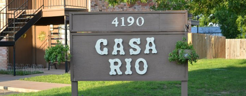Sign reading '4190 CASA RIO' in front of an apartment building with exterior stairs and landscaping.