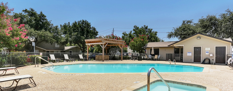 Community pool with lounge chairs and a pergola, flanked by apartment buildings and trees, on a sunny day.