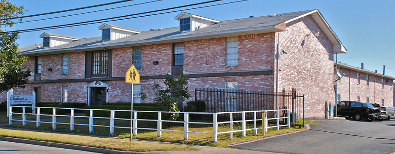 Two-story brick apartment building with white fencing and a pedestrian crossing sign, under a clear sky.