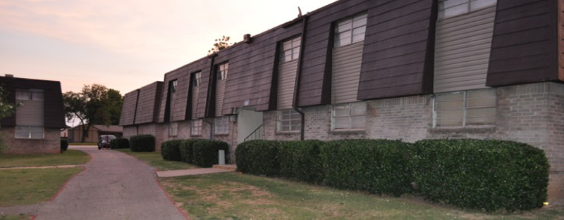 Long, two-story apartment building with dark shutters, a hedged walkway, and a fading evening sky.