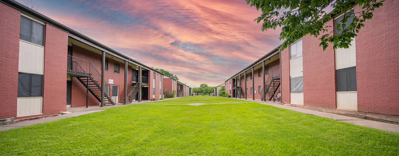 Symmetrical view of a grassy courtyard flanked by red-bricked, two-story apartment buildings with external stairs, under a dramatic pink and orange sunset sky.