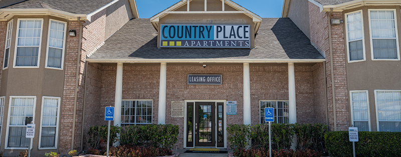 Entrance of 'COUNTRY PLACE APARTMENTS' with a 'LEASING OFFICE' sign, handicapped parking signage, and neatly trimmed bushes under a clear sky.