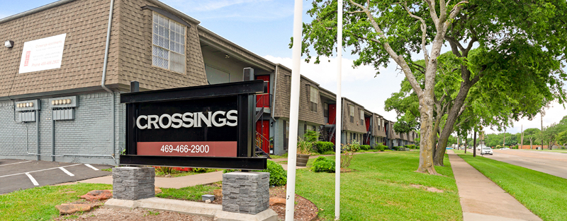 'CROSSINGS' signboard with contact details in front of a brick building on the left, and a sidewalk beside a grassy area with trees lining a street next to apartment buildings on the right.