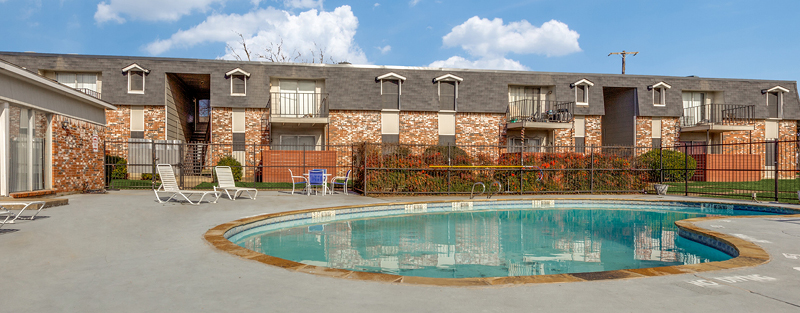 Outdoor swimming pool with lounge chairs in the foreground and a two-story apartment building with brick exterior and balcony railings in the background, under a partly cloudy sky.
