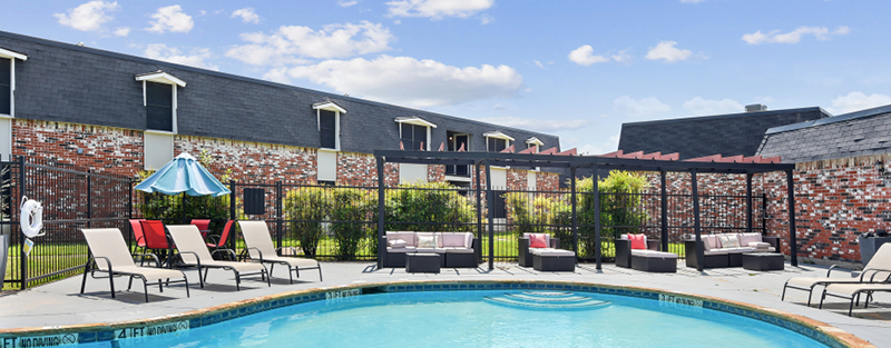 Outdoor swimming pool with lounge chairs and patio furniture, enclosed by an iron fence, with a two-story apartment complex in the background.
