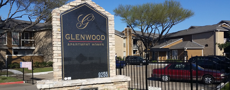 Entrance sign for 'Glenwood Apartment Homes' with a stone pedestal and background of apartment buildings and parked cars.