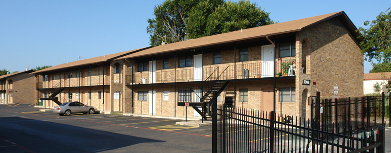 Two-story brown brick apartment building with balconies and external staircases, enclosed by a black iron fence, with a parking lot in the foreground.