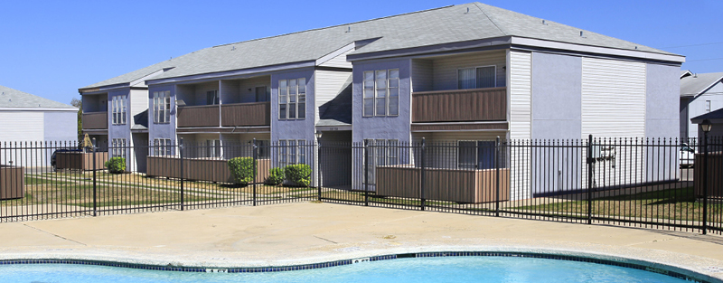 Swimming pool in the foreground with a two-story apartment building behind a metal fence under a clear blue sky.