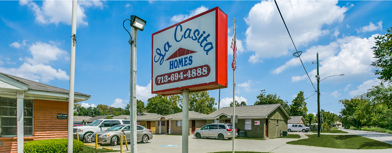 A bright sign for 'La Casita HOMES' with a phone number, next to a parking lot and small single-story buildings under a blue sky with clouds.