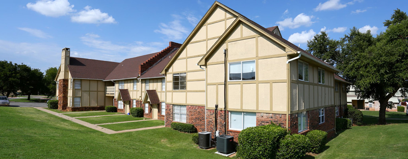 A beige two-story apartment building with brick accents and a pitched roof, set on a well-manicured lawn under a blue sky with scattered clouds.