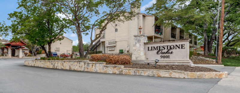 A stone sign for 'LIMESTONE Oaks 9001' in front of the garden-style two-story apartment complex with lush trees and a clear sky.
