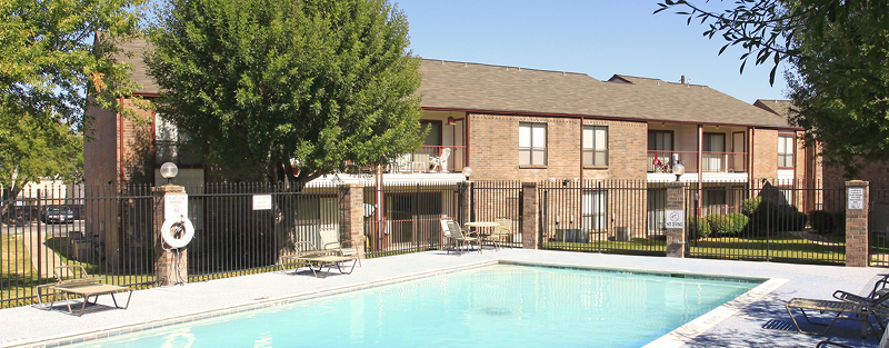 Outdoor swimming pool with clear blue water, surrounded by a metal fence, with two-story brick apartment buildings and trees in the background on a sunny day.