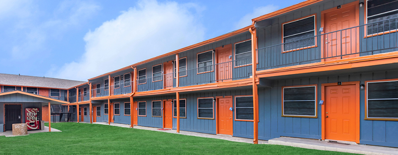 Two-story apartment complex with orange doors and trim, grey siding, and a central courtyard, under a cloudy sky.