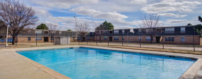 A large outdoor swimming pool in the foreground with a fenced apartment complex and bare trees in the background on a cloudy day.