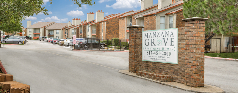 Entrance of Manzana Grove Apartment Homes with a brick sign, a gated parking lot, and two-story buildings with red trim roofs.