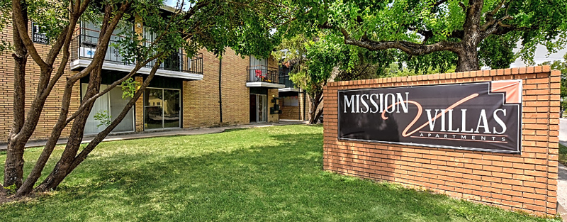 Large sign reading 'MISSION VILLAS APARTMENTS' in front of a brick building with balconies, shaded by mature trees on a lush green lawn.