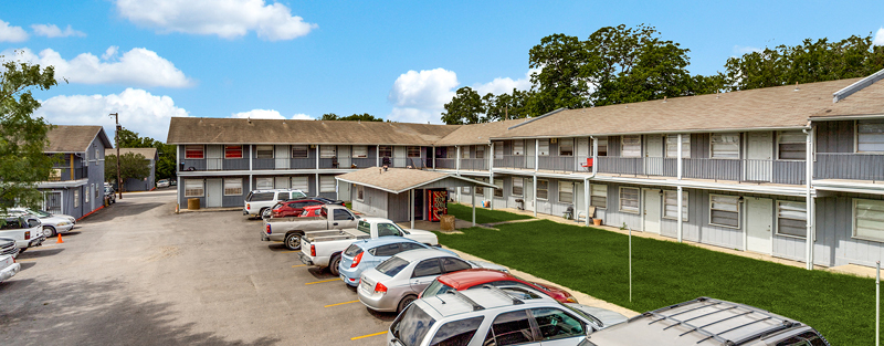 Two-story residential apartment complex with parked cars in the lot, featuring gray siding, on a sunny day with scattered clouds.