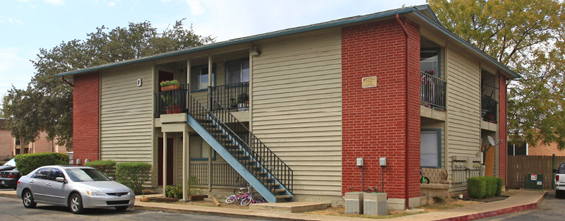 A two-story apartment complex with beige siding and brick red exterior, blue stairs, parked cars, and a child's bike in the foreground.