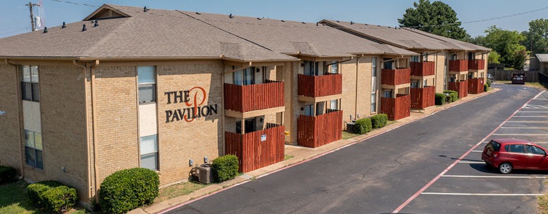 A two-story brick apartment building with red balcony accents and a sign reading 'THE PAVILION,' alongside a parking lot with a red car.