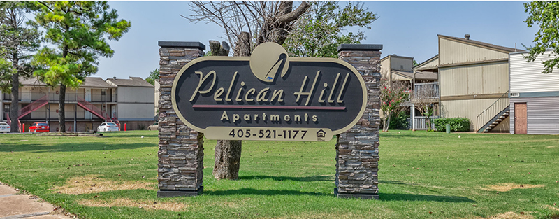 Signage for 'Pelican Hill Apartments' with contact information, set against a backdrop of apartment buildings and green lawn.