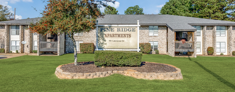 Large sign reading 'PINE RIDGE APARTMENTS' in a well-manicured lawn in front of a brick apartment complex with balconies and vibrant trees.