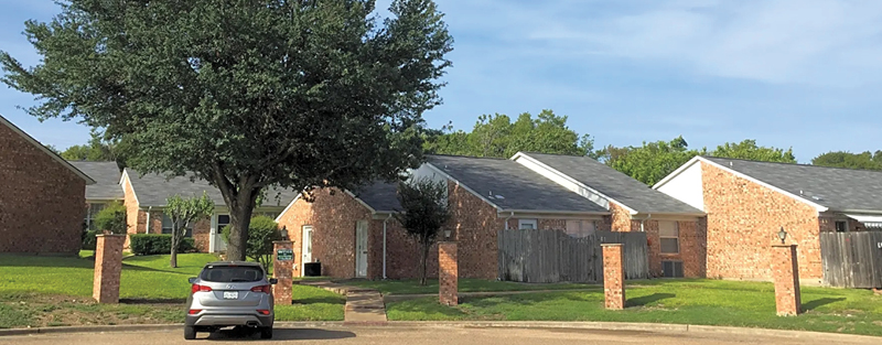 Single-story residential brick homes with a central large tree and a parked car on a sunny day.