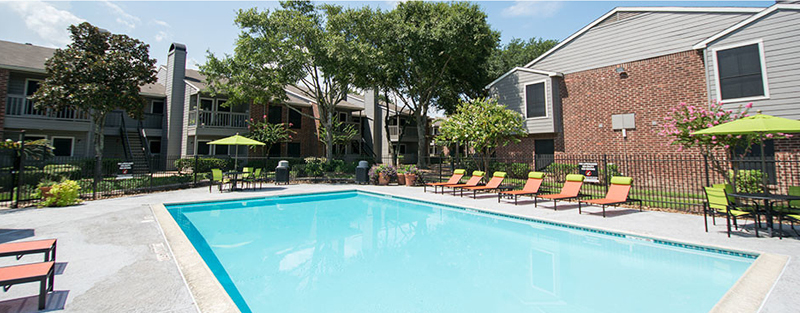 A clear blue swimming pool with surrounding lounge chairs and patio umbrellas in an apartment complex courtyard with brick buildings and green trees.