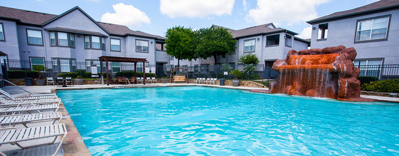 Swimming pool with a faux rock waterfall feature, surrounded by lounge chairs and a gray two-story apartment complex, under a partly cloudy sky.