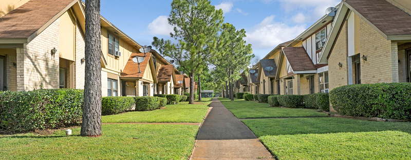 Pathway leading through a landscaped lawn between rows of two-story townhomes with pointed roofs under a clear sky.