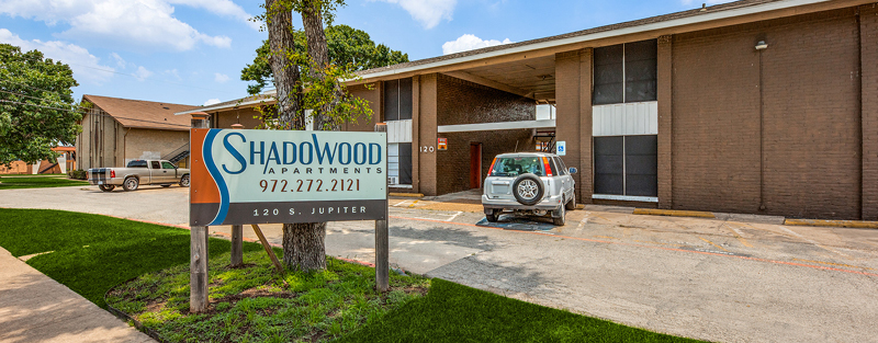 Entrance of 'SHADOWOOD APARTMENTS' with a signboard and contact number, next to a brown brick building with parked cars under a partly cloudy blue sky.