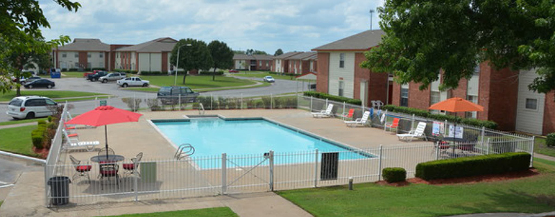 Community swimming pool with red umbrellas and lounge chairs, enclosed by a metal fence, with red brick apartment buildings in the background, on a cloudy day.