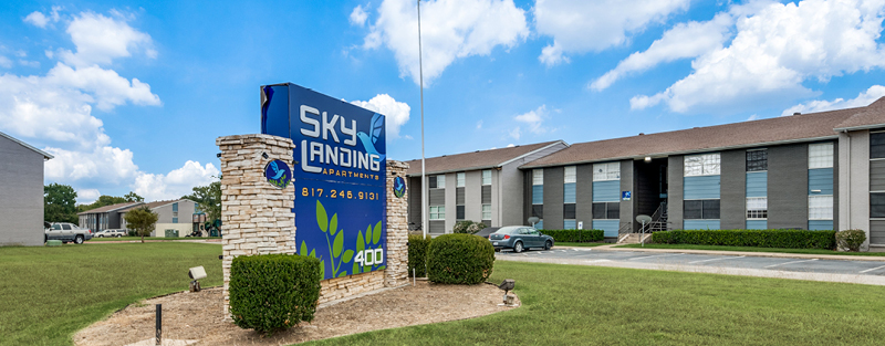 A prominent 'SKY LANDING APARTMENTS' sign in blue with a contact number, in front of a two-story residential building with parked cars and partly cloudy skies.