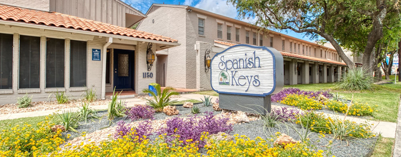 "Spanish Keys apartment complex sign surrounded by colorful flowerbeds, with a two-story building in the background.