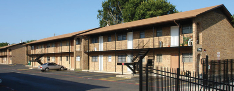 Two-story brown brick apartment complex with external metal staircases, parking lot, and a black wrought iron fence, in bright daylight.