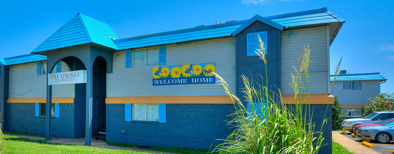 A blue two-story apartment building with two signs, one saying, 'THE SPRINGS OF MOORE,' and another, 'WELCOME HOME,' along with tall grass in the foreground under a blue sky.