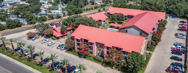 Aerial view of a Stepping Stone Apartments, with red roofs, lush landscaping, and a full parking lot, on a bright day.