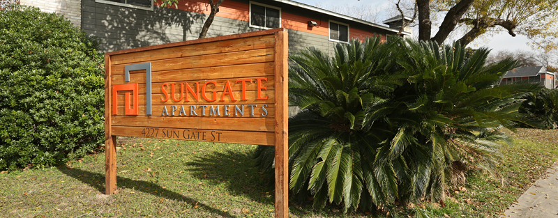 Wooden signboard for Sungate Apartments next to a small palm tree, in front of a multicolored apartment building.
