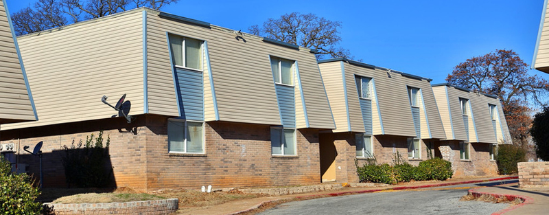 Tan siding two-story apartment building with blue trim and satellite dishes, brick lower walls, and a clear blue sky.