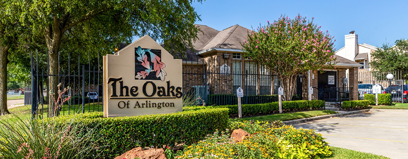 Sign for 'The Oaks Of Arlington' in front of the gated apartment community with landscaped beds and a background of trees and buildings on a sunny day.